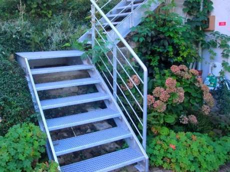 A steel grating stair outside the house is surrounded by flowers and trees.